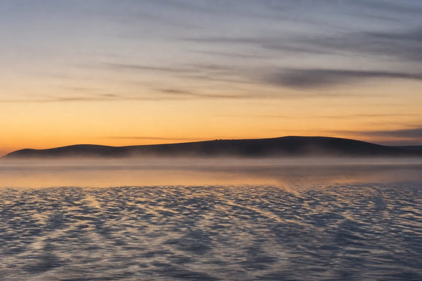 Mist lying flat on still water at first light, with a low dark headland along the horizon and ribbed wet sand in the foreground.